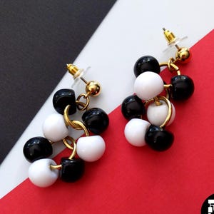Pode incluir: Um par de brincos pendurados com aglomerados de contas pretas e brancas. Os brincos são conectados com aros e pinos de metal dourado. O fundo é uma divisão diagonal de vermelho, branco e preto.