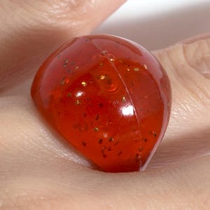 May include: A chunky, translucent orange ring featuring a smooth, rounded surface embedded with small, reflective gold-colored glitter particles throughout the material. The ring has a visible seam running vertically along the center of the band.
