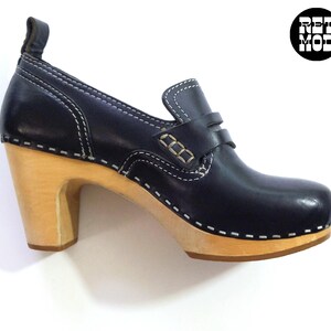 May include: A black leather clog-style shoe with a wooden heel. The shoe features white stitching details and a small strap across the top. The heel is approximately 3 inches high. The shoe has a classic, vintage aesthetic.