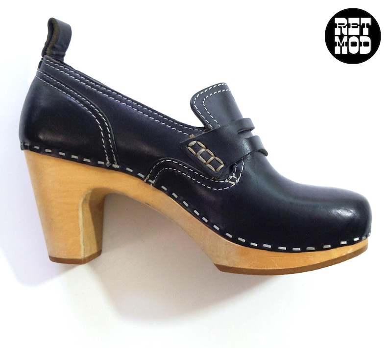 May include: A black leather clog-style shoe with a wooden heel. The shoe features white stitching details and a small strap across the top. The heel is approximately 3 inches high. The shoe has a classic, vintage aesthetic.