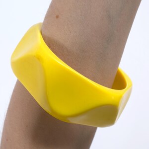 May include: A bright yellow bangle bracelet with a smooth, glossy finish and a distinct geometric, faceted design featuring multiple angled surfaces that create a bold, sculptural appearance around the circumference of the circular wrist accessory.