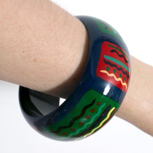 May include: A wide, dark blue bangle bracelet featuring a repeating pattern of rectangular blocks in red and green. Each block is decorated with horizontal wavy lines and dots in yellow, black, and light green, creating a vibrant, colorful design.