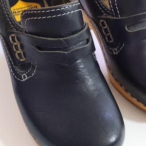 May include: Black leather clogs with a wooden sole and white stitching. The clogs have a strap across the top and are labeled "Offel, Made in Sweden". These shoes are a classic style.