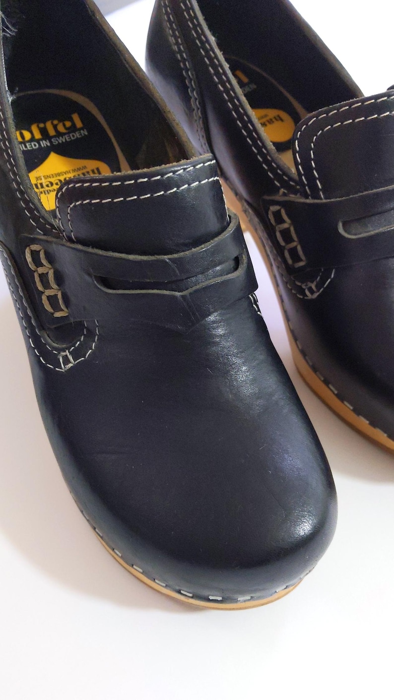 May include: Black leather clogs with a wooden sole and white stitching. The clogs have a strap across the top and are labeled "Offel, Made in Sweden". These shoes are a classic style.