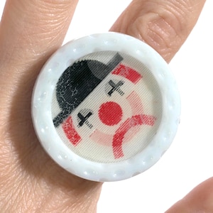 May include: A white ring featuring a circular bottle cap design. The center graphic displays a clown face with a black hat, a red nose, and red mouth details. The ring has a textured, scalloped edge resembling a standard metal bottle cap.