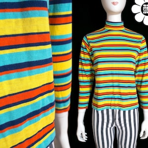 May include: A long-sleeve mock neck shirt featuring horizontal stripes in vibrant shades of yellow, turquoise, orange, and navy blue. The knit fabric displays a consistent pattern across the torso and sleeves, ending in a simple hemline.
