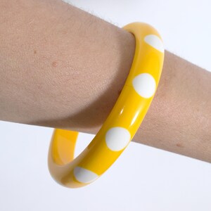 May include: A bright yellow bangle bracelet featuring a repeating pattern of solid white circular polka dots. The accessory has a smooth, glossy finish and a rounded, thick band construction that encircles the wrist with a vibrant color contrast.