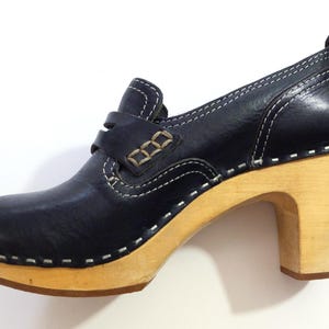 May include: A navy blue leather clog shoe with a wooden heel. The shoe features white stitching, a strap across the top, and a pull tab at the back. The heel measures approximately 3 inches.
