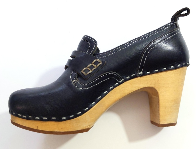 May include: A navy blue leather clog shoe with a wooden heel. The shoe features white stitching, a strap across the top, and a pull tab at the back. The heel measures approximately 3 inches.