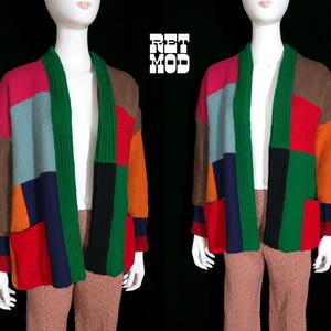 May include: A color-blocked knit cardigan featuring a patchwork design in shades of red, green, blue, orange, brown, and pink. The garment has a ribbed green collar, long sleeves, and two front pockets. The text RET MOD is visible in white.