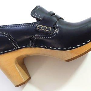 May include: A navy blue leather clog with a light wood heel and sole. The shoe features white stitching and a strap across the top. The heel is approximately 3 inches high.