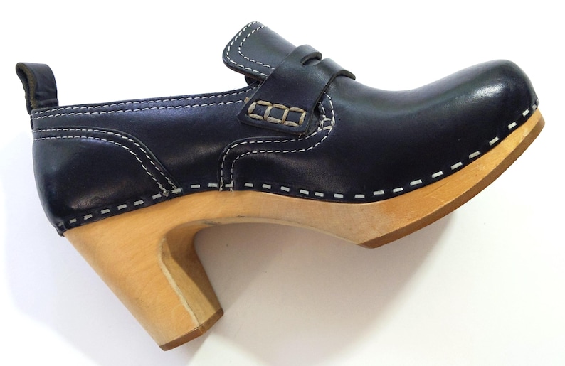 May include: A navy blue leather clog with a light wood heel and sole. The shoe features white stitching and a strap across the top. The heel is approximately 3 inches high.