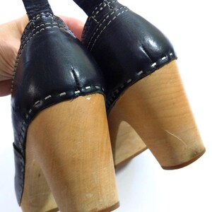 May include: A pair of black leather clogs with wooden heels. The black leather uppers are stitched with white thread. The wooden heels are a light brown color.