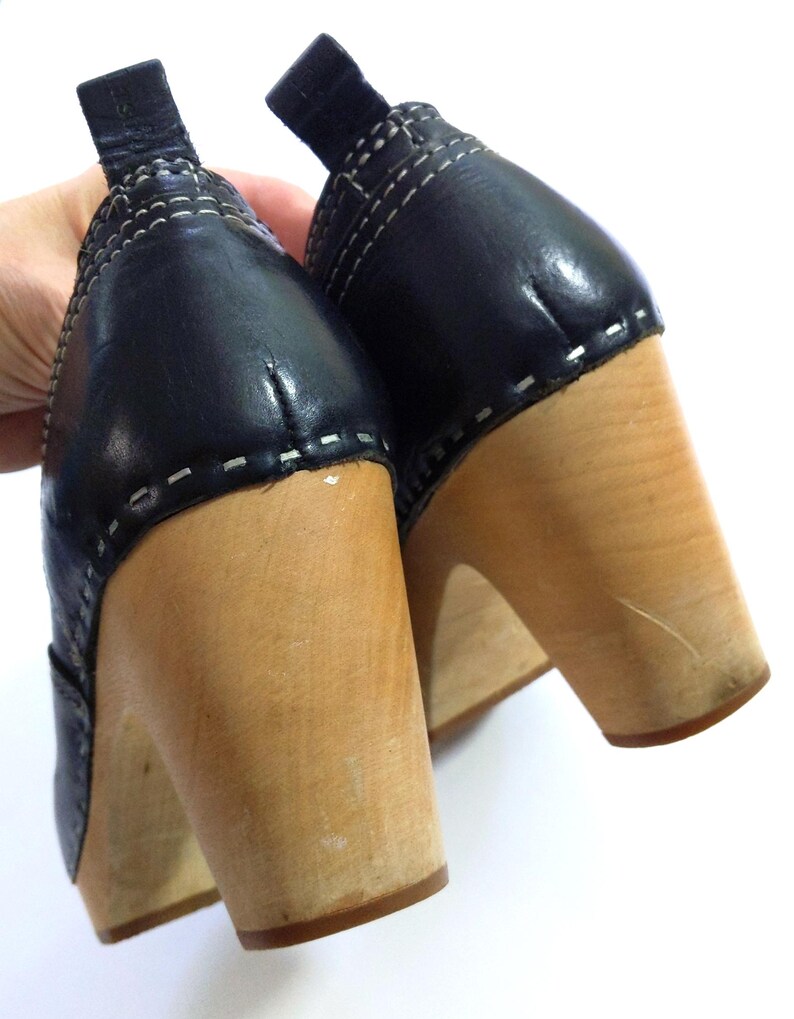 May include: A pair of black leather clogs with wooden heels. The black leather uppers are stitched with white thread. The wooden heels are a light brown color.