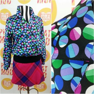 Könnte beinhalten: Eine schwarze und blaue gemusterte Bluse mit Kragen und langen Ärmeln. Die Bluse hat ein geometrisches Muster aus Kreisen mit verschiedenen farbigen Segmenten. Die Bluse wird mit einem rosa und schwarzen karierten Rock kombiniert.