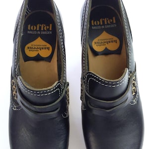 May include: Pair of black leather clogs with white stitching and gold-colored accents. The clogs have a pull tab at the heel and the brand name "toffel" is visible on the insole, along with "NAILED IN SWEDEN" and "hasbeens".