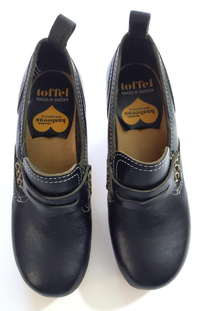 May include: Pair of black leather clogs with white stitching and gold-colored accents. The clogs have a pull tab at the heel and the brand name "toffel" is visible on the insole, along with "NAILED IN SWEDEN" and "hasbeens".
