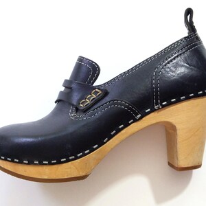 May include: Black leather clog-style shoe with a wooden heel. The shoe features a rounded toe, a strap across the top with gold-colored accents, and white stitching details. The heel is approximately 3 inches high.