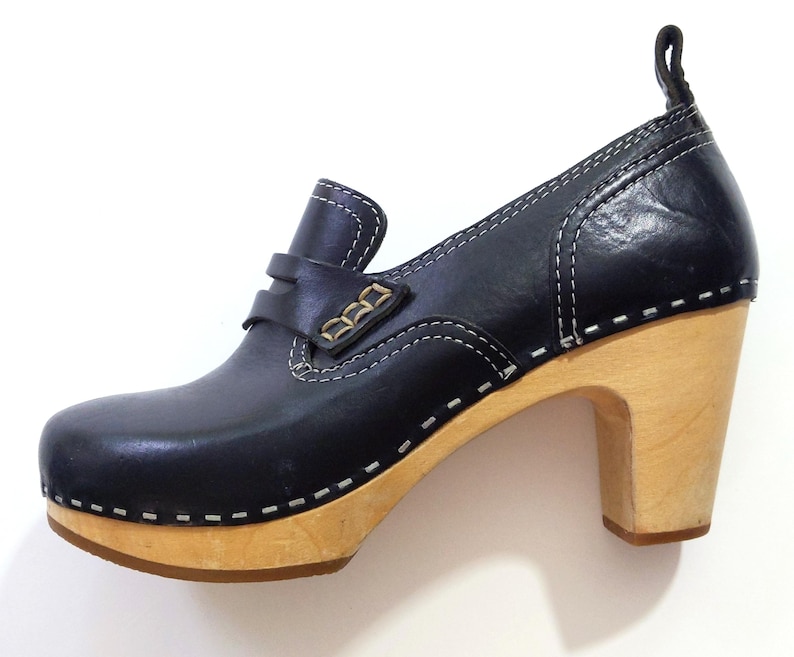 May include: Black leather clog-style shoe with a wooden heel. The shoe features a rounded toe, a strap across the top with gold-colored accents, and white stitching details. The heel is approximately 3 inches high.