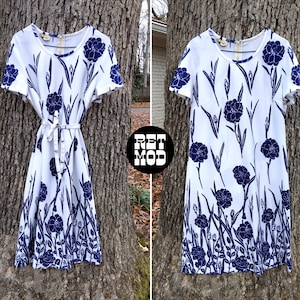 May include: White dress with a navy blue floral pattern. The dress has short sleeves, a round neckline, and a tie belt. The floral design includes large flowers and stems. The dress is hanging on a tree.