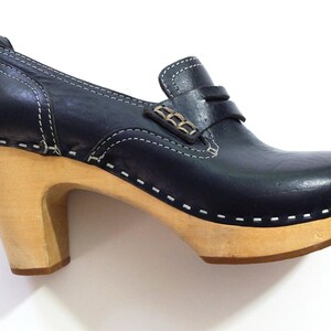 May include: A navy blue clog shoe with a light wood heel and sole. The shoe features a leather upper with white stitching and a decorative strap with metal accents. The heel is approximately 3 inches high.