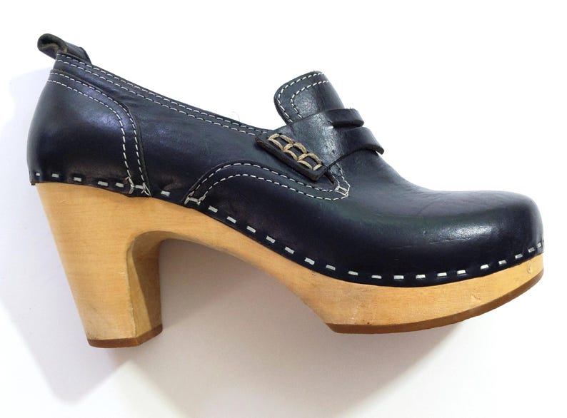 May include: A navy blue clog shoe with a light wood heel and sole. The shoe features a leather upper with white stitching and a decorative strap with metal accents. The heel is approximately 3 inches high.