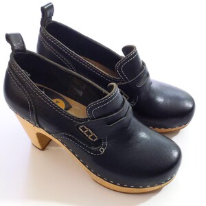 May include: A pair of black leather clogs with a wooden sole and a heel. The shoes have white stitching and a pull tab at the back. The heels are approximately 3 inches tall.
