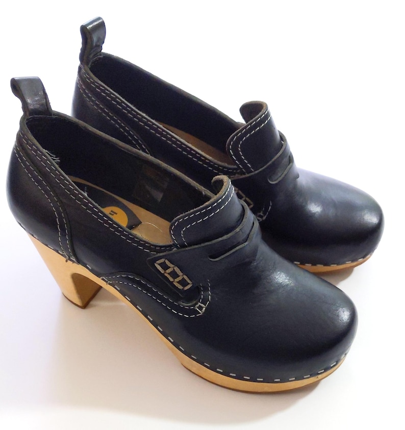 May include: A pair of black leather clogs with a wooden sole and a heel. The shoes have white stitching and a pull tab at the back. The heels are approximately 3 inches tall.
