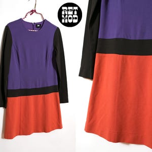 May include: A color block dress with long black sleeves, a purple bodice, a black band, and a red-orange skirt. The dress is hanging on a hanger against a white background. The dress has a small "W" logo.