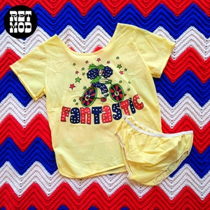 May include: A yellow vintage pajama set featuring a short-sleeved top and matching bottoms. The top has a graphic of a woman riding a bicycle with the word "FANTASTIC" in colorful letters. The set is displayed on a red, white, and blue patterned blanket.