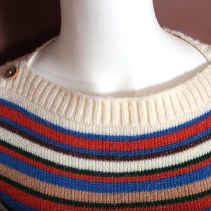 May include: A striped sweater with a cream-colored neckline and shoulder detail. The sweater features horizontal stripes in red, blue, brown, and white. Wooden buttons adorn the shoulders.