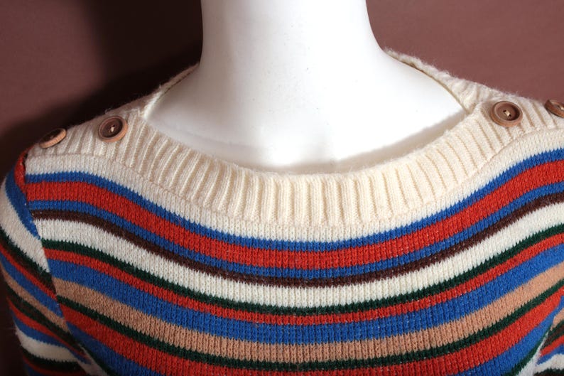 May include: A striped sweater with a cream-colored neckline and shoulder detail. The sweater features horizontal stripes in red, blue, brown, and white. Wooden buttons adorn the shoulders.