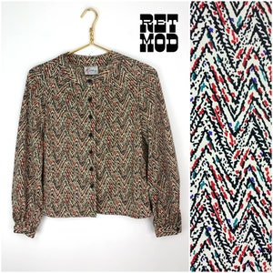 May include: A beige, red, black and white patterned button-up blouse with long sleeves. The blouse has a mandarin collar and a chevron pattern.