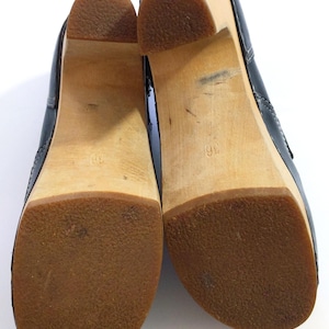 May include: Pair of black clogs with wooden soles and brown rubber bottoms. The shoes have a classic design with a rounded toe and a slight heel. The soles are marked with the number 36.