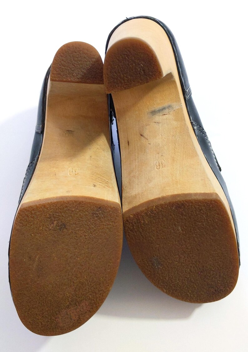 May include: Pair of black clogs with wooden soles and brown rubber bottoms. The shoes have a classic design with a rounded toe and a slight heel. The soles are marked with the number 36.