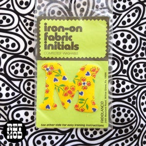 May include: A yellow fabric letter M with a floral and heart pattern in red, blue, and green, packaged in a clear plastic sleeve with a lime green card backing labeled iron-on fabric initials and completely washable by Freelance.
