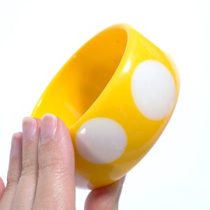 May include: A bright yellow bangle bracelet featuring a smooth, glossy finish and a repeating pattern of large, solid white polka dots. The circular accessory has a thick, rounded profile and a wide band design that is uniform in its vibrant color.