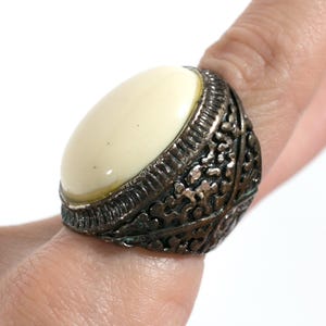 May include: A ring featuring a large, smooth, oval-shaped cream-colored stone set in a dark, oxidized metal band. The band displays an intricate, textured, and perforated pattern across its surface, providing a detailed contrast to the stone.