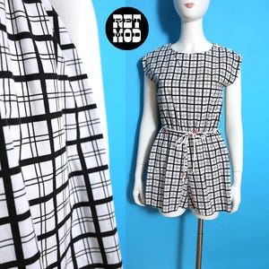 Puede incluir: Un mono a cuadros blanco y negro con cuello redondo y mangas cortas. El mono tiene un cinturón blanco y detalles de botones rojos. El maniquí está sobre un fondo azul brillante. El logotipo "RET MOD" está en un círculo negro.