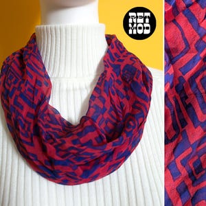 Könnte beinhalten: Ein roter und blauer Schal mit Muster. Der Schal hat ein sich wiederholendes geometrisches Design in Rot mit blauen Akzenten. Der Schal ist um einen weißen Rollkragenpullover drapiert.