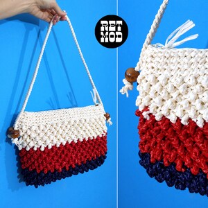 May include: A white, red, and blue crocheted shoulder bag with wooden bead accents. The bag has a long strap and is made of a textured yarn.