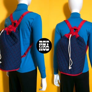 Può includere: Uno zaino trapuntato blu navy con finiture e spalline rosse. La chiusura con coulisse è bianca. Lo zaino è esposto su un manichino con un dolcevita blu. Il logo "RET MOD" è visibile.