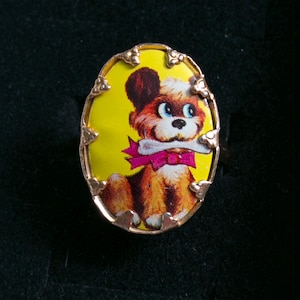 May include: An oval-shaped ring featuring a gold-toned metal setting with decorative prongs. The center inlay displays a vibrant yellow background with a colorful illustration of a brown puppy holding a white bone and wearing a bright pink bow.