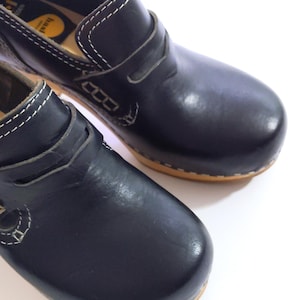 May include: Pair of black leather clogs with a wooden sole. The clogs have a rounded toe and a strap across the top. White stitching accents the edges and details. The shoes are set against a white background.