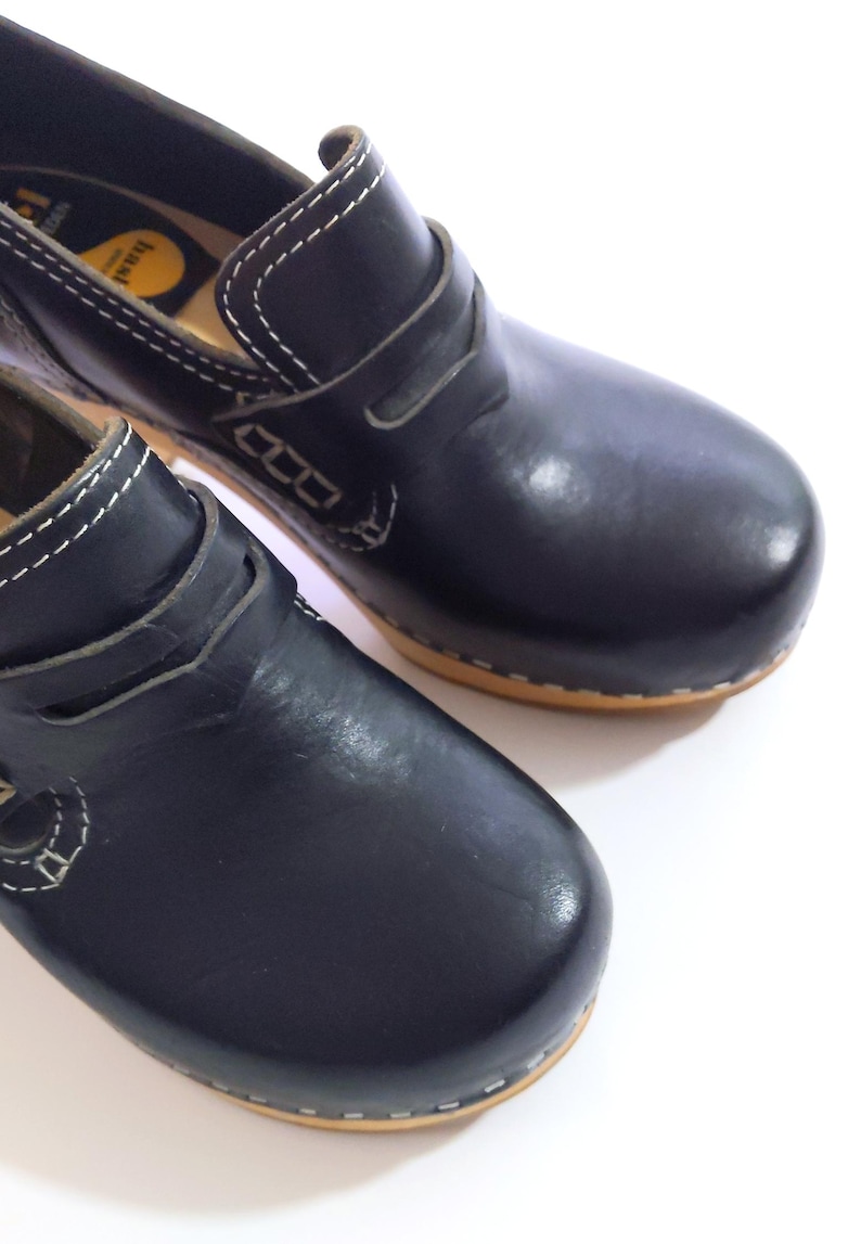 May include: Pair of black leather clogs with a wooden sole. The clogs have a rounded toe and a strap across the top. White stitching accents the edges and details. The shoes are set against a white background.