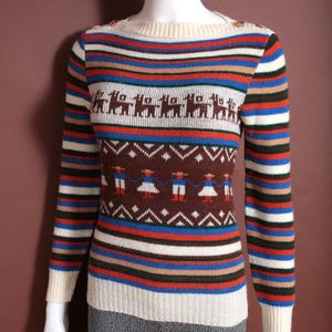 May include: A vintage-style sweater with a striped pattern in cream, blue, red, and brown. The sweater features a crew neck and long sleeves. The design includes brown llamas and figures with geometric patterns. The sweater is displayed on a mannequin.