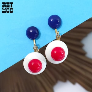 May include: A pair of retro earrings. Each earring has a white circular base with a red centre and a navy blue sphere at the top. The earrings are suspended from gold-coloured clasps. The image includes the text "RET MOD".