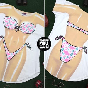 May include: A white t-shirt featuring a front and back graphic print of a tan torso wearing a pink and white bikini with a flamingo and leaf pattern. The front shows the bikini top and bottom, while the back shows the bikini straps and bottom.