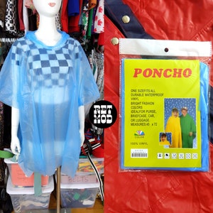 May include: A blue plastic poncho with a checkered pattern. The poncho is packaged in a clear plastic bag with a yellow label that reads "PONCHO" and includes details about the product, such as size, material, and color. The label also includes a rainbow graphic and the text "100% VINYL".
