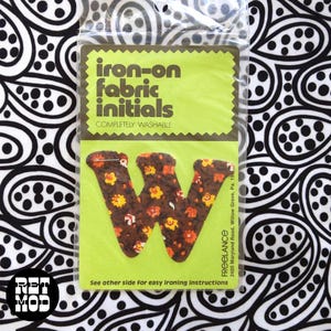 May include: A brown letter W iron-on fabric initial patch with a floral pattern of small orange and yellow flowers. The patch is packaged in a clear plastic sleeve with a lime green card featuring the text iron-on fabric initials.